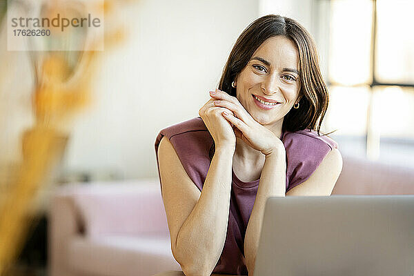 Lächelnde Geschäftsfrau mit der Hand am Kinn sitzt zu Hause