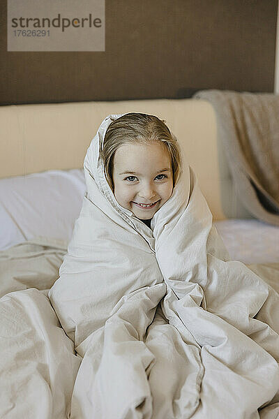 Lächelndes süßes Mädchen  eingewickelt in eine Decke  das zu Hause auf dem Bett sitzt