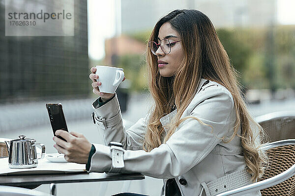 Frau hält Kaffeetasse mit Smartphone im Straßencafé in der Hand