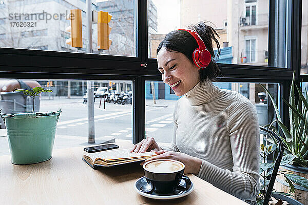 Glückliche Frau  die im Café sitzt und Lieder über drahtlose Kopfhörer hört und ein Buch liest