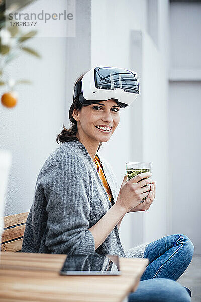 Lächelnde Geschäftsfrau im Virtual-Reality-Simulator mit Trinkglas auf Stuhl sitzend