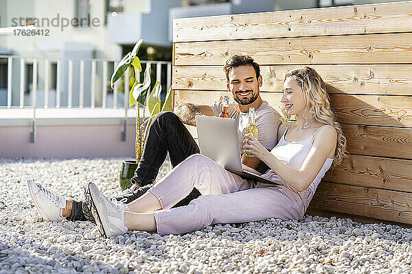 Lächelndes junges Paar  das an einem sonnigen Tag mit einem Laptop sitzt und Bier anstößt