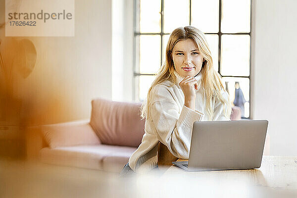 Lächelnde blonde Geschäftsfrau sitzt mit Laptop zu Hause