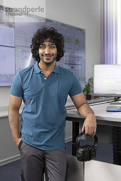 Lächelnder Techniker steht mit der Hand in der Tasche am Schreibtisch