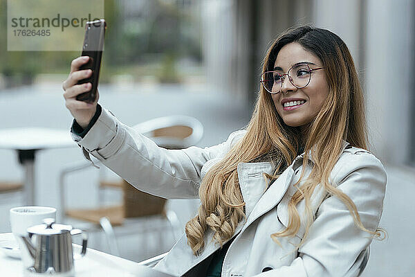 Lächelnde Frau  die im Straßencafé sitzt und ein Selfie mit dem Handy macht