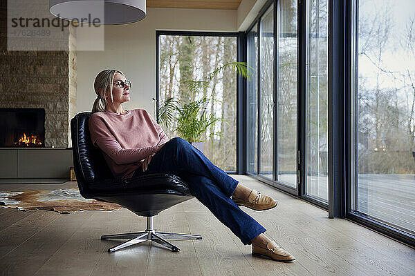Frau schaut durch Glasfenster und sitzt auf einem Stuhl im Wohnzimmer