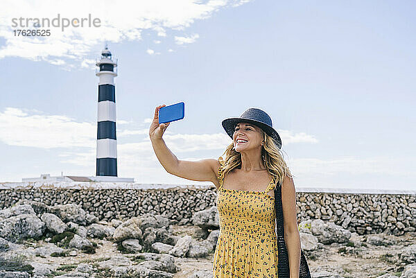 Happy mature woman taking selfie through smart phone at Artrutx Lighthouse  Minorca  Spain