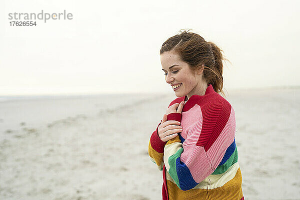 Glückliche junge Frau in buntem Pullover steht am Strand