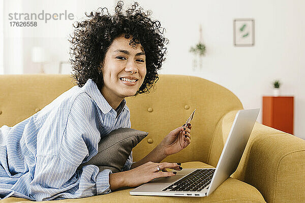 Glückliche junge Frau mit Kreditkarte und Laptop  die zu Hause auf dem Sofa liegt