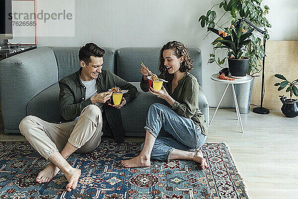 Glückliches junges Paar mit Essen zum Mitnehmen  das zu Hause auf dem Sofa im Wohnzimmer sitzt