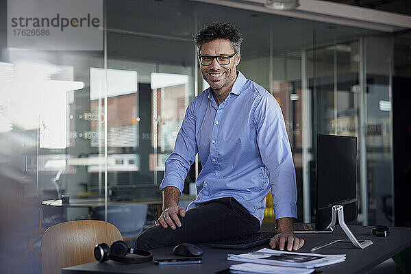 Glücklicher Geschäftsmann mit Brille sitzt am Schreibtisch am Arbeitsplatz