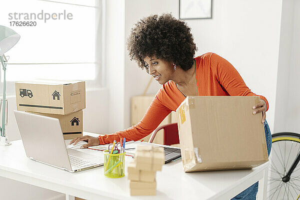 Lächelnder Freiberufler mit Paket und Laptop  der am Tisch steht und von zu Hause aus arbeitet