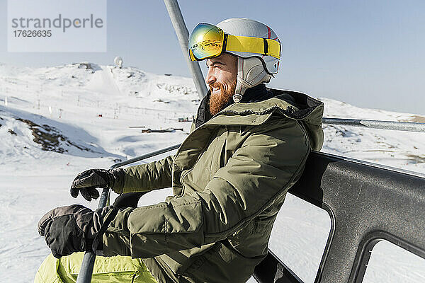 Lächelnder junger Mann mit Jacke  Skibrille und Helm sitzt am Skilift