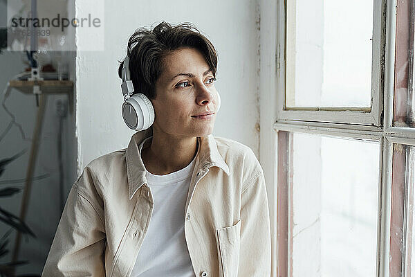 Nachdenkliche Frau mit Kopfhörern  die am Fenster Musik hört