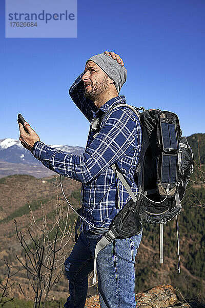Mann mit Rucksack und Smartphone  das an einem sonnigen Tag über ein tragbares Solarpanel aufgeladen wird