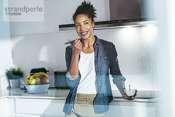 Lächelnde Frau  die in der Küche steht und über einen Lautsprecher mit ihrem Mobiltelefon telefoniert