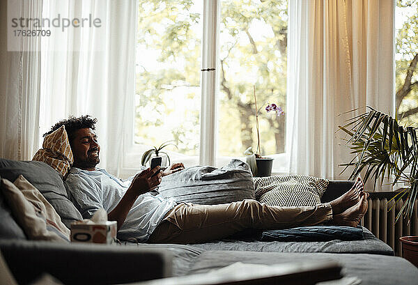 Smiling man surfing net through smart phone lying on sofa in living room