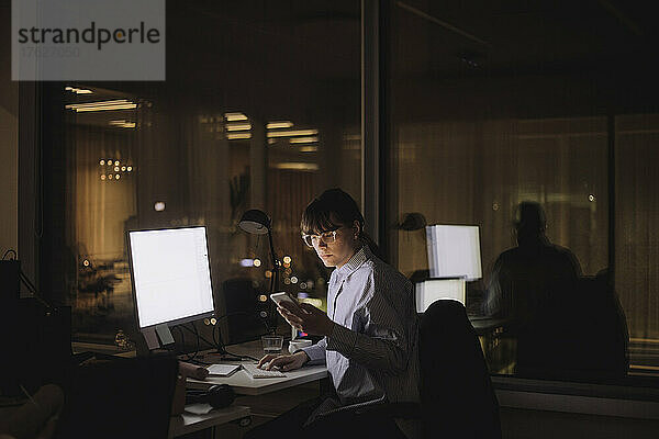 Geschäftsfrau  die nachts im Büro sitzend eine Textnachricht auf ihrem Mobiltelefon schreibt