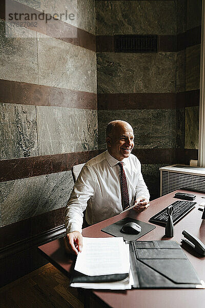Lächelnd arbeitender älterer männlicher Finanzberater  der mit einem Dokument in einer Anwaltskanzlei sitzt