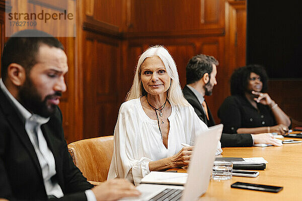 Porträt einer lächelnden Geschäftsfrau  die neben einem Geschäftsmann sitzt  der einen Laptop am Konferenztisch im Sitzungssaal benutzt