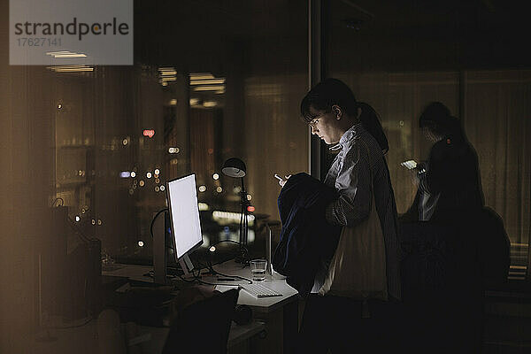 Eine Geschäftsfrau benutzt ihr Smartphone  während sie nachts im Büro arbeitet