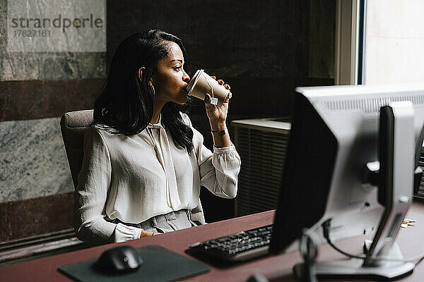 Weibliche Anwältin trinkt Kaffee aus einem Einwegbecher  während sie am Schreibtisch im Büro sitzt