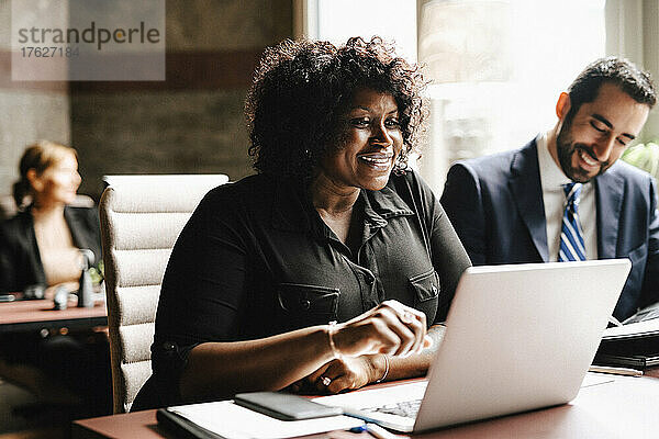 Lächelnde Geschäftsfrau  die einen Laptop benutzt  während ein Geschäftsmann am Schreibtisch sitzt und in einer Anwaltskanzlei arbeitet