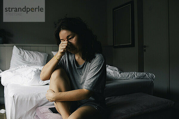 Traurige Frau weint  während sie auf der Bank am Bettende im Schlafzimmer zu Hause sitzt