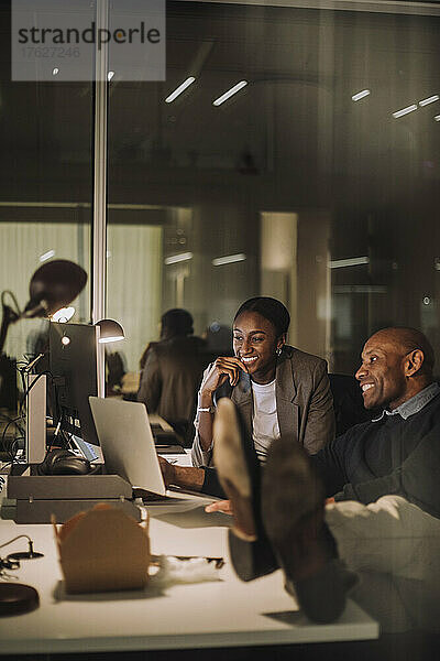 Lächelnde männliche und weibliche Kollegen  die über einen Laptop diskutieren  während sie in letzter Minute nachts im Büro arbeiten