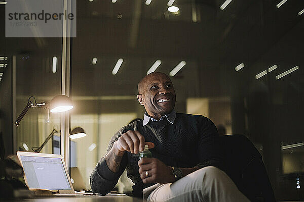 Glücklicher Geschäftsmann mit Saftflasche im Büro sitzend bei Nacht