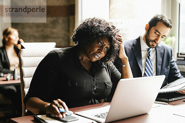 Geschäftsfrau mit Hand in den Haaren  die einen Laptop benutzt  während ein Geschäftsmann im Büro arbeitet