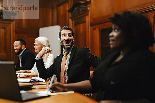 Glücklicher Geschäftsmann mit offenem Mund  der inmitten von Kollegen in einer Konferenzsitzung im Sitzungssaal sitzt