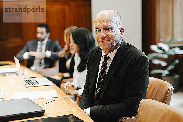 Porträt eines lächelnden älteren männlichen Anwalts im Sitzungssaal mit Kollegen während einer Sitzung