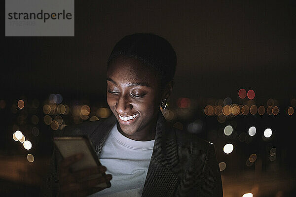 Glückliche Geschäftsfrau  die nachts eine SMS auf ihrem Smartphone schreibt