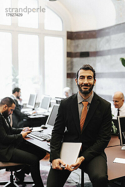 Porträt eines lächelnden männlichen Anwalts  der einen Laptop hält und auf einem Schreibtisch im Büro sitzt
