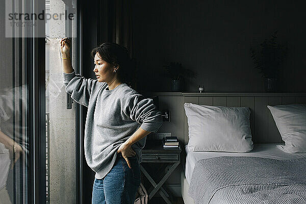 Nachdenkliche einsame Frau  die aus dem Fenster schaut  während sie mit der Hand auf der Hüfte im Schlafzimmer zu Hause steht