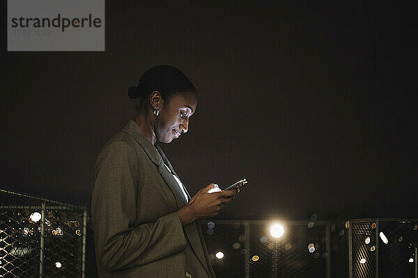 Seitenansicht einer Geschäftsfrau  die nachts eine Textnachricht auf einem Mobiltelefon schreibt