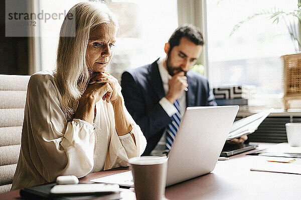 Reife Geschäftsfrau starrt auf Laptop von Geschäftsmann am Schreibtisch im Büro
