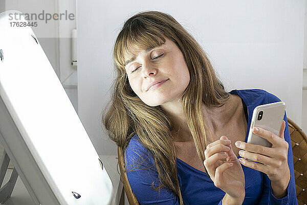 Frau sitzt mit ihrem Smartphone neben einer Lichttherapielampe.