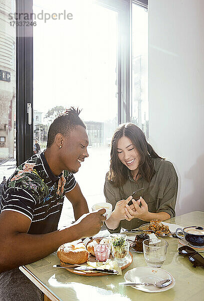 Lächelndes Paar genießt das Frühstück und schaut im Restaurant auf das Smartphone