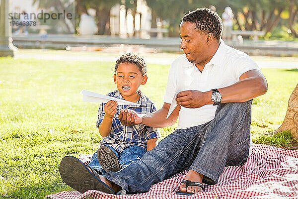 Glücklicher afroamerikanischer Vater und gemischtrassiger Sohn spielen mit Papierflugzeugen im Park