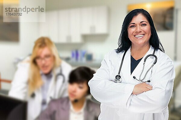 Hübsche hispanische Ärztin oder Krankenschwester  die in ihrem Büro steht und deren Mitarbeiter dahinter arbeiten