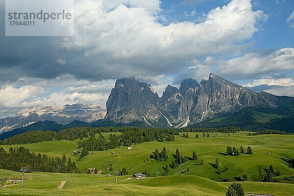 In Südtirol auf der Seiser Alm im Frühsommer mit einigen Almhütten und einem Schilift auf saftig grünen Weiden. Im Hintergrund ist ein großer felsiger Berg mit einigen Wolken am blauem Himmel