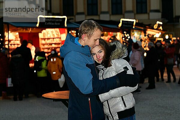 Vor den beleuchteten Verkaufsständen an einem Weihnachtsmarkt steht ein junges verliebtes Paar und hält sich im Arm