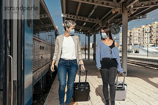 Two happy female friends in protective masks walking and chatting on the platform at train station