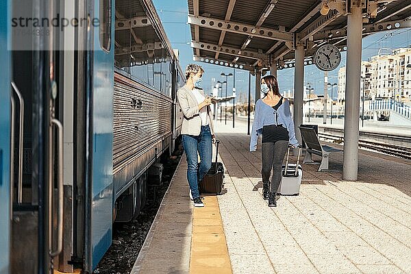 Two happy female friends in protective masks walking and chatting on the platform at train station