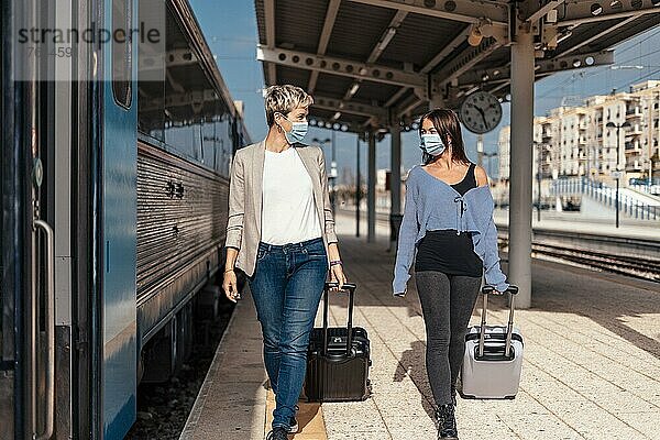 Two happy female friends in protective masks walking and chatting on the platform at train station