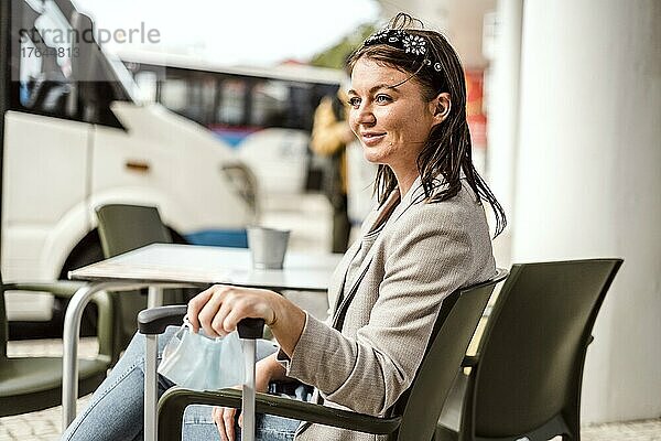 A young traveler with the luggage and protective mask sitting in the cafe and waiting for her bus to come