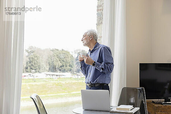 Älterer Mann mit Kaffeetasse  der durch das Fenster schaut und im Hotelapartment steht