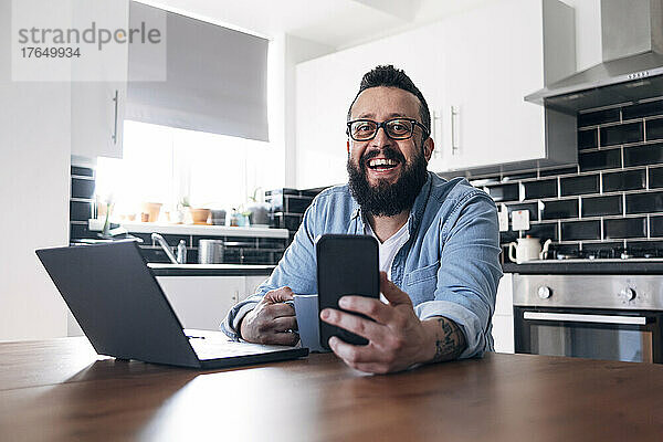 Glücklicher Freiberufler mit Laptop und Smartphone  der zu Hause in der Küche sitzt